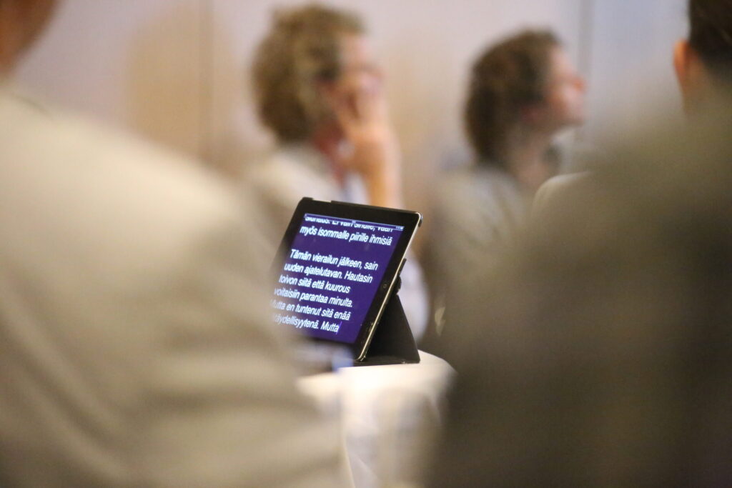Eine Gruppe die gespannt einem Vorttrag folgen mit Blick auf ein Tablet der Schriftdolmetscher*inneb