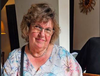 Frau mit kurzen, lockigen, graubraunen Haaren und Brille trägt eine floral gemusterte Bluse und eine Halskette,