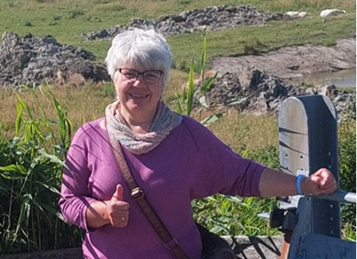 Frau mit kurzen weißen Haaren, trägt lila Pullover und Daumen hoch, steht im Freien vor grüner Landschaft