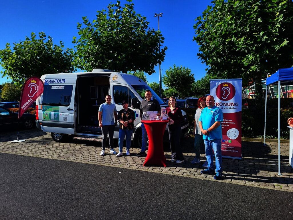 Ein mobiler Informationsstand mit einem weißen Fahrzeug im Hintergrund. Fünf Personen stehen um einen roten Tisch, auf dem Informationsmaterial liegt. Im Hintergrund sind grüne Bäume sichtbar.