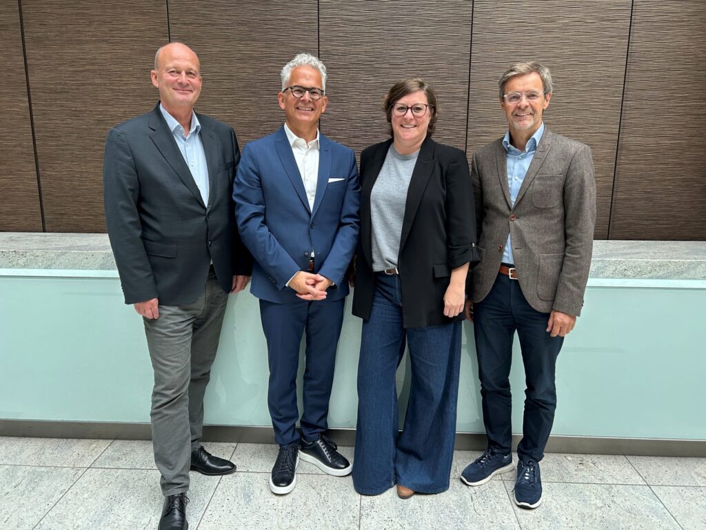 Vier Personen stehen nebeneinander vor einer Wand aus Holzpaneelen. Eine Person trägt einen blauen Anzug, die anderen sind in formeller Kleidung. Die Frau in der Mitte trägt eine schwarze Jacke und weite Jeans.