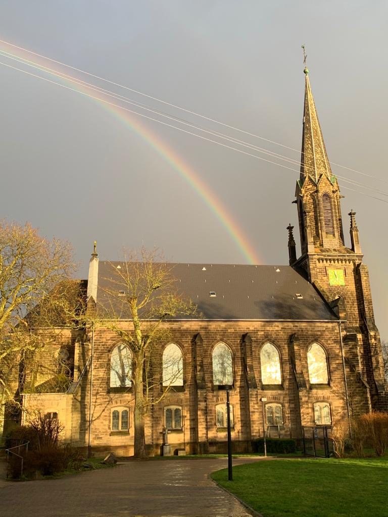 Kirche mit spitzem Turm und Regenbogen am Himmel darüber, Bäume ohne Blätter im Vordergrund