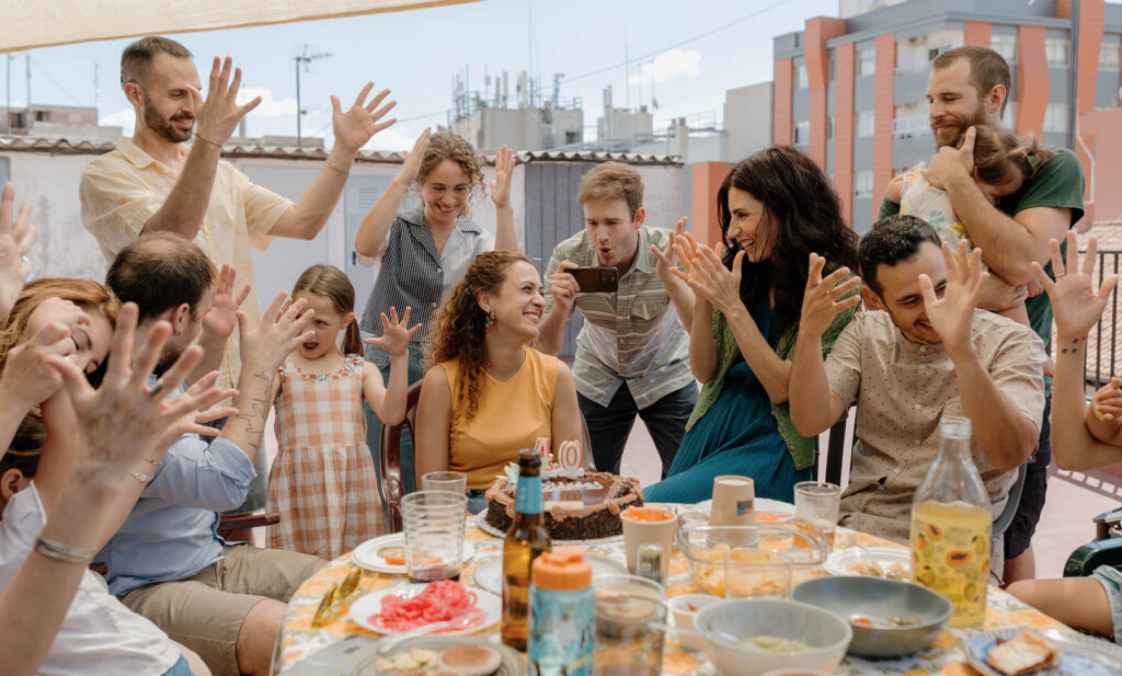 Menschen sitzen und stehen um einen gedeckten Tisch auf einer Dachterrasse, heben die Hände und feiern mit einem Kuchen mit brennender Kerze.
