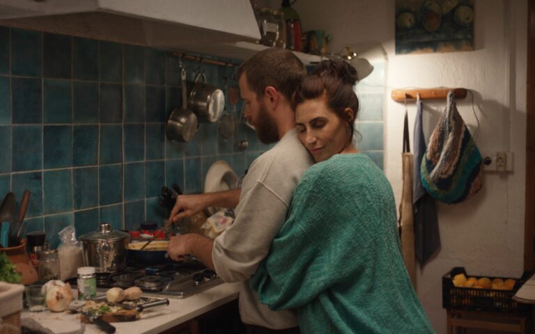 Zwei Personen in einer Küche, eine steht vor dem Herd und kocht, die andere umarmt sie von hinten.