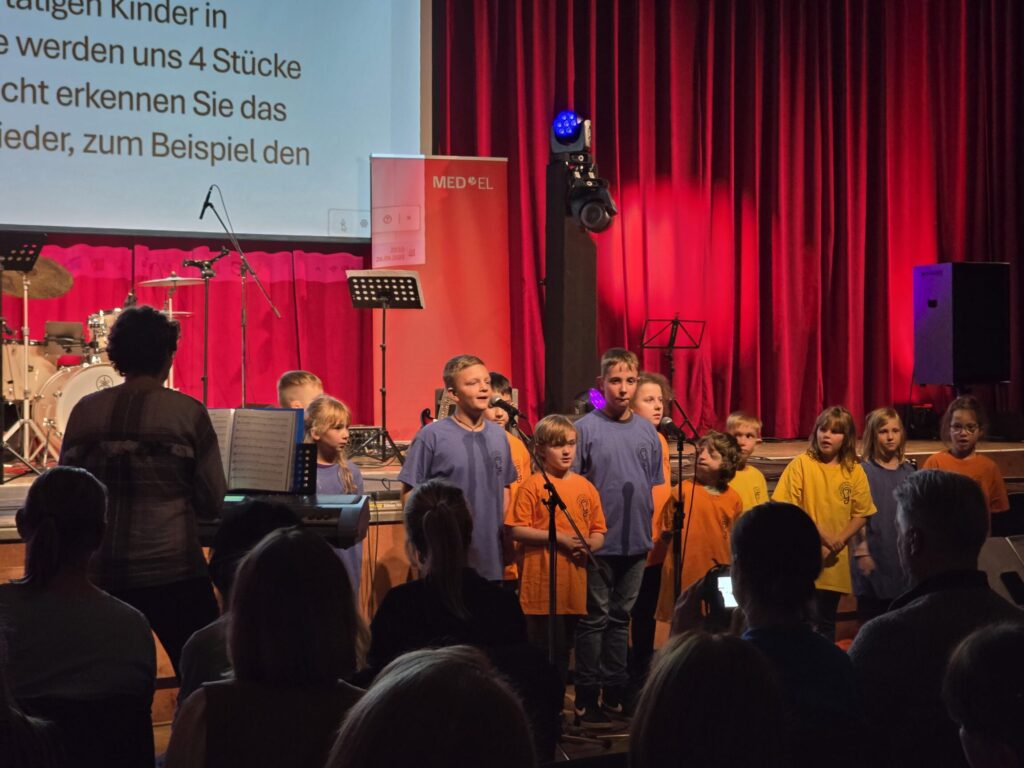 Kinder stehen auf einer Bühne vor Mikrofonen, einige tragen blaue, andere gelbe T-Shirts, im Hintergrund roter Vorhang und Musik-Equipment.
