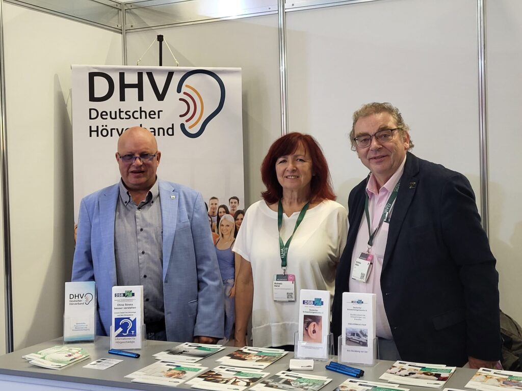 Messestand des Deutschen Schwerhörigenbundes mit drei Personen hinter Infotresen, Informationsmaterial und Bannern mit Logos und Texten zu Hörbehinderung