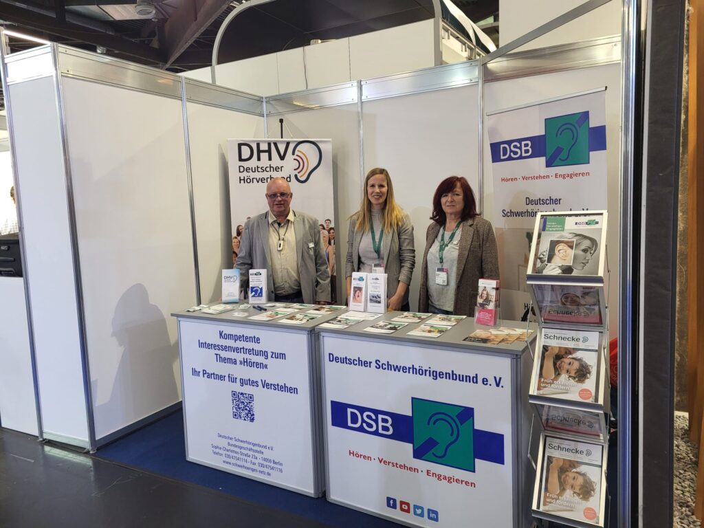 Messestand des Deutschen Schwerhörigenbundes mit drei Personen hinter Infotresen, Informationsmaterial und Bannern mit Logos und Texten zu Hörbehinderung