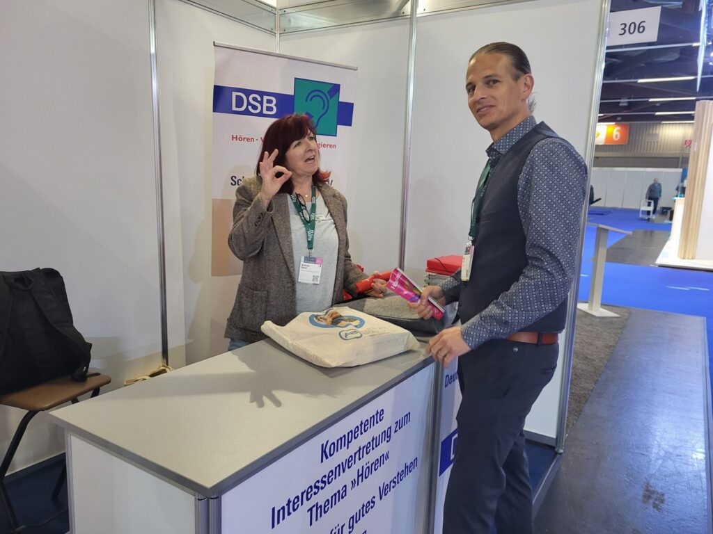 Messestand mit Banner des DSB, zwei Personen im Gespräch, eine zeigt ein Handzeichen, auf dem Tisch liegt eine Tasche.