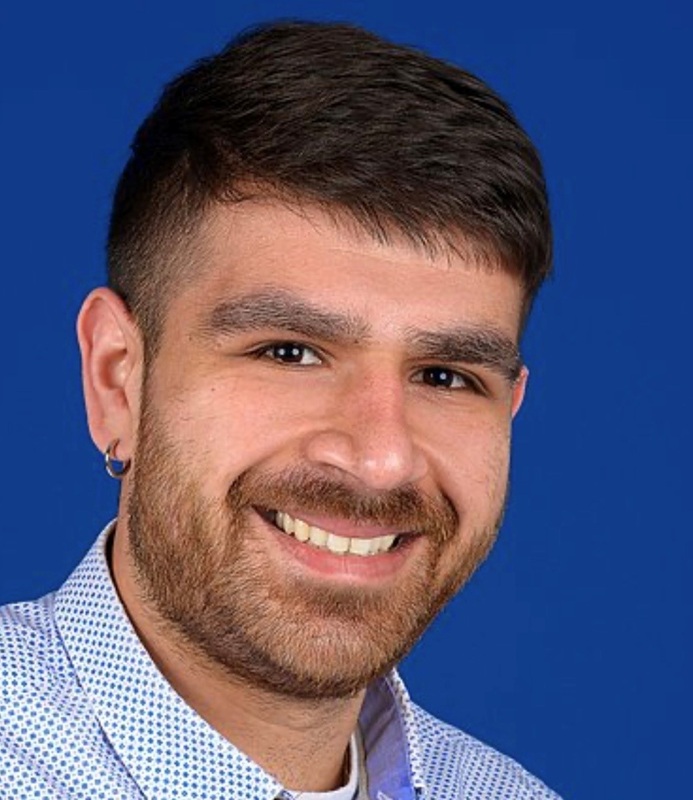 Mann mit kurzem, dunklem Haar, Hemd mit blauem Punktmuster und silbernem Ohrstecker vor blauem Hintergrund.