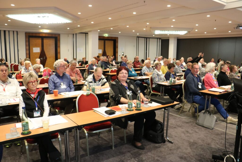 Teilnehmer sitzen in einem Konferenzraum mit Reihen von Tischen und Stühlen. Auf den Tischen stehen Wasserflaschen und Schreibmaterial.