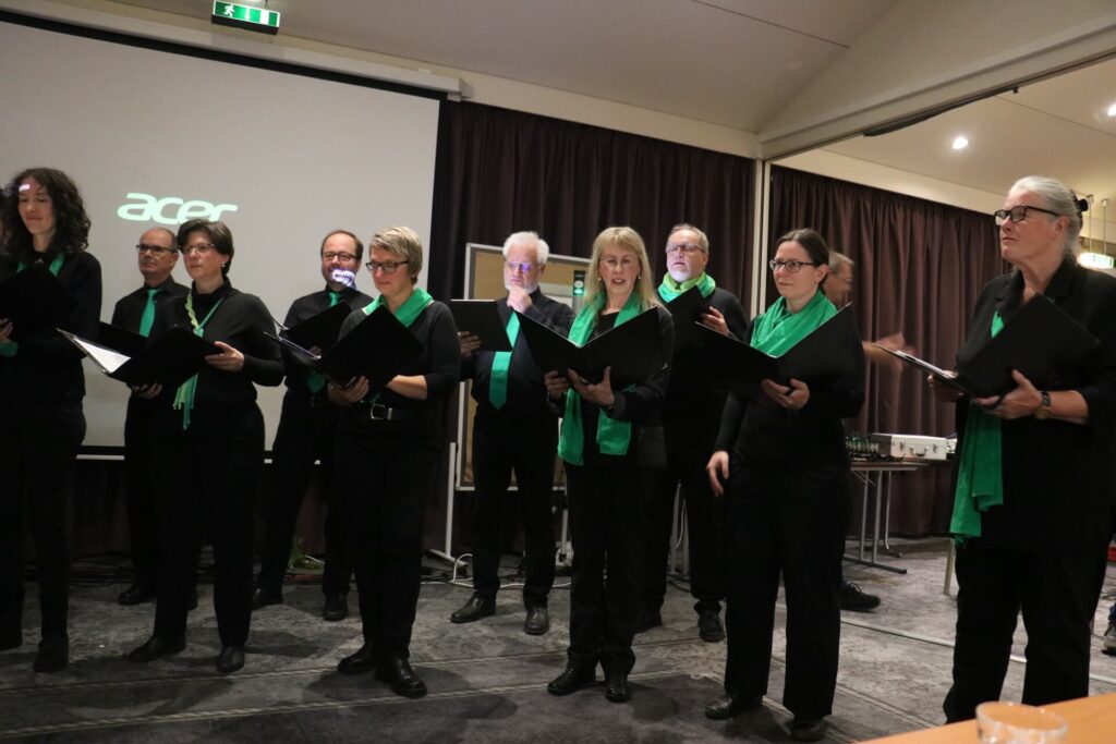 Chemnitzer Kammerchor steht in einem Raum, trägt schwarze Kleidung mit grünen Schals, hält schwarze Ordner, im Hintergrund eine Leinwand.