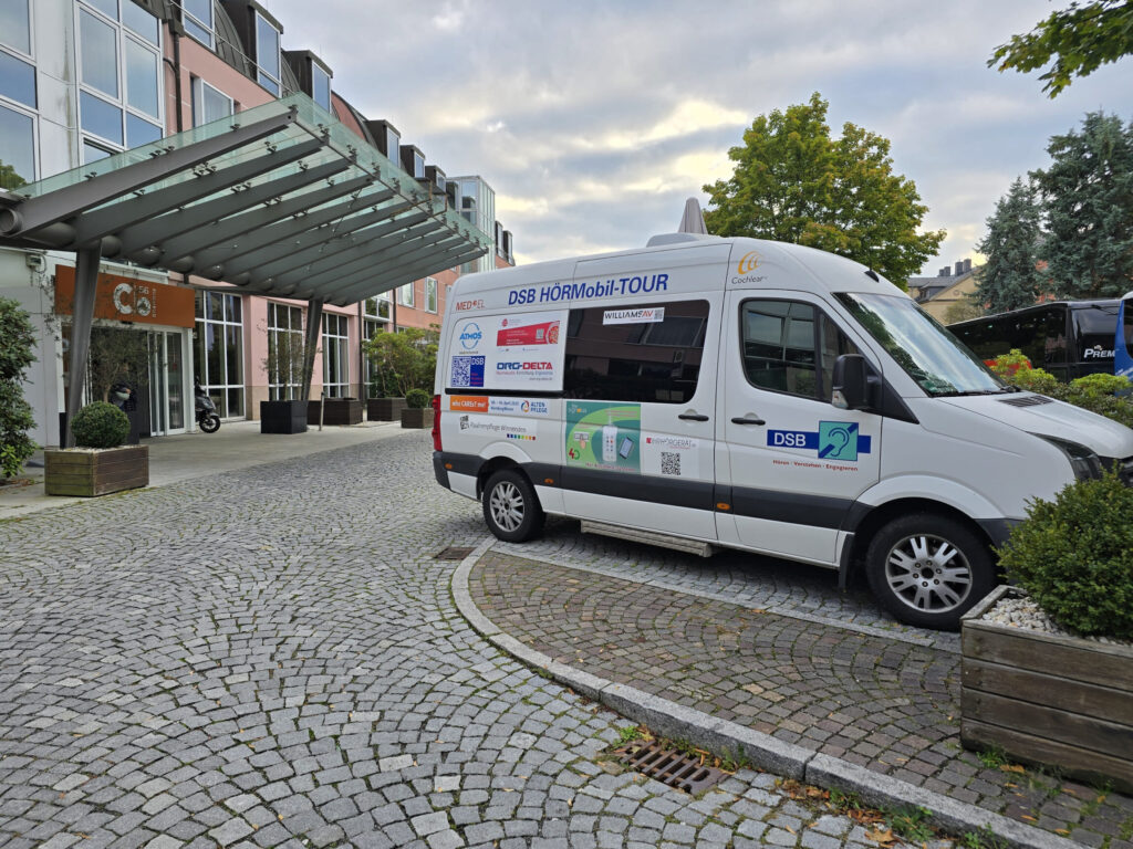 Weißer Transporter mit Aufschrift 'DSB HÖRMobil-TOUR' und verschiedenen Logos steht vor einem Gebäude mit Glasvordach auf gepflastertem Hof.