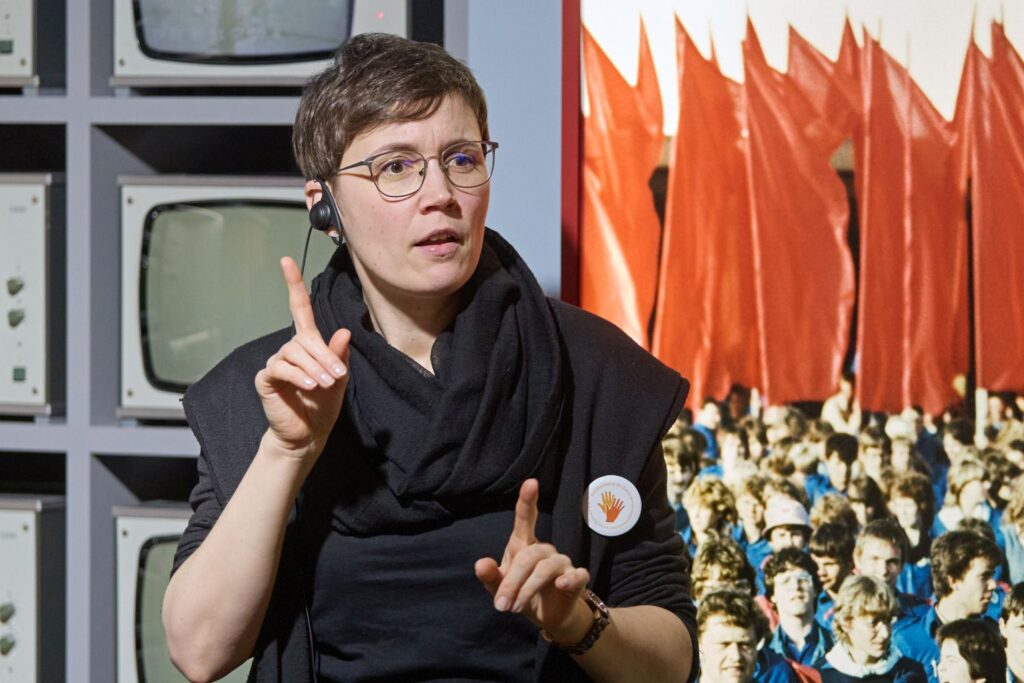 Person mit schwarzem Schal und Headset zeigt mit beiden Händen nach oben.