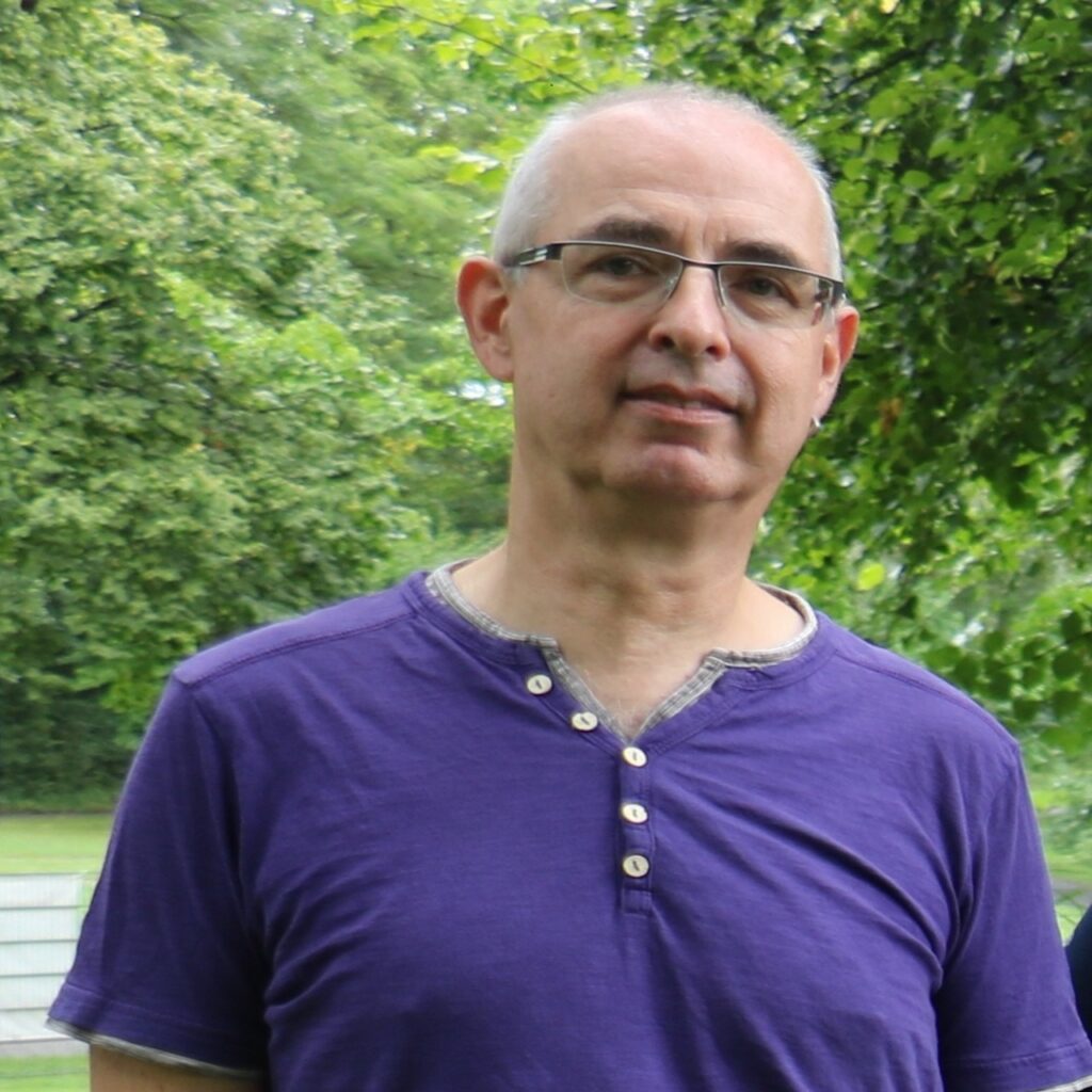 Mann mit Brille und lila T-Shirt mit Knopfleiste steht vor grünen Bäumen im Freien