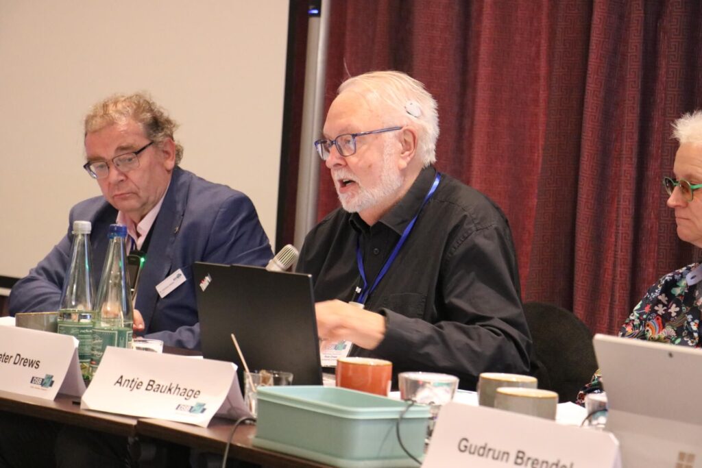 Drei Personen sitzen an einem Konferenztisch mit Mikrofonen, Laptops und Namensschildern vor einem roten Vorhang.