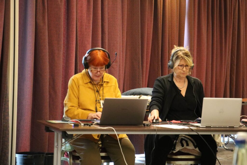 Zwei Personen mit Headsets sitzen an einem Tisch mit zwei geöffneten Laptops vor einem rotbraunen Vorhang.