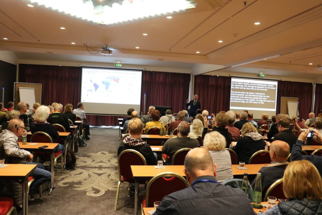 Konferenzraum mit Publikum, das einem Vortrag folgt; vorne zwei Projektionsflächen mit Landkarte und Text.