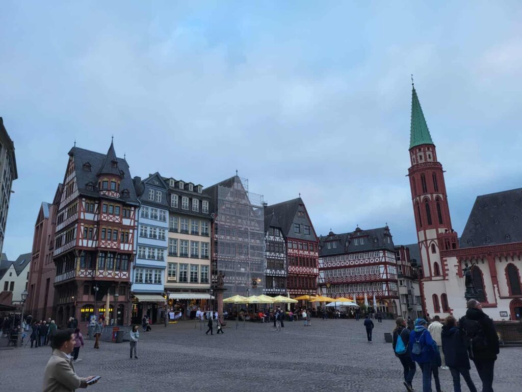 Historische Fachwerkhäuser und ein Kirchturm mit grünem Dach an einem großen Platz bei bewölktem Himmel