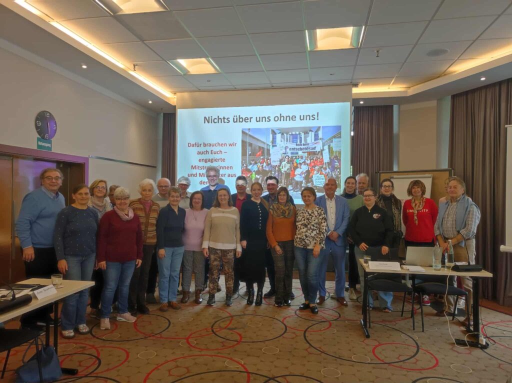 Gruppe von Menschen steht in einem Tagungsraum vor einer Leinwand mit Text und Bild, umgeben von Tischen und Stühlen.