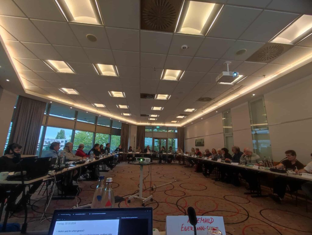 Konferenzraum mit U-förmig angeordneten Tischen, an denen Personen sitzen, Deckenbeleuchtung und Fenster mit Blick ins Freie.