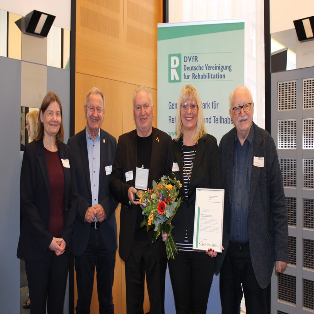 Fünf Personen stehen nebeneinander, eine hält einen Blumenstrauß und eine Urkunde, im Hintergrund ein Banner der Deutschen Vereinigung für Rehabilitation.