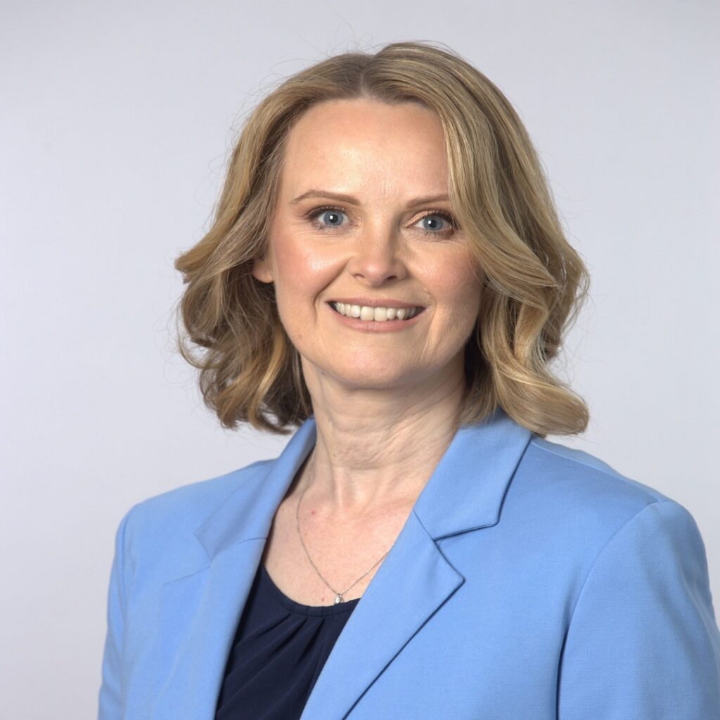 Frau mit schulterlangem, gewelltem blonden Haar trägt hellblauen Blazer und dunkles Oberteil, steht vor hellem Hintergrund.