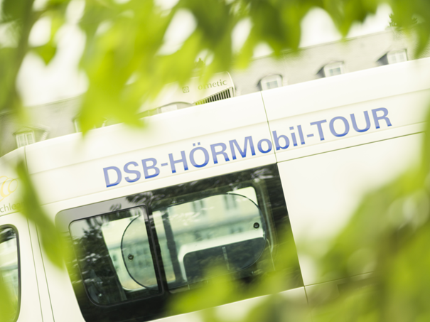 Teilansicht eines weißen Fahrzeugs mit der blauen Aufschrift 'DSB HÖRMobil-TOUR' hinter grünen Blättern.