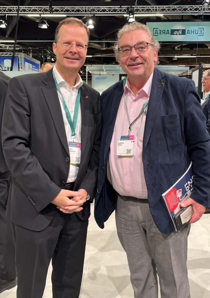 Zwei Männer in Anzügen mit Namensschildern auf einer Messe, einer hält eine Mappe und ein Smartphone.