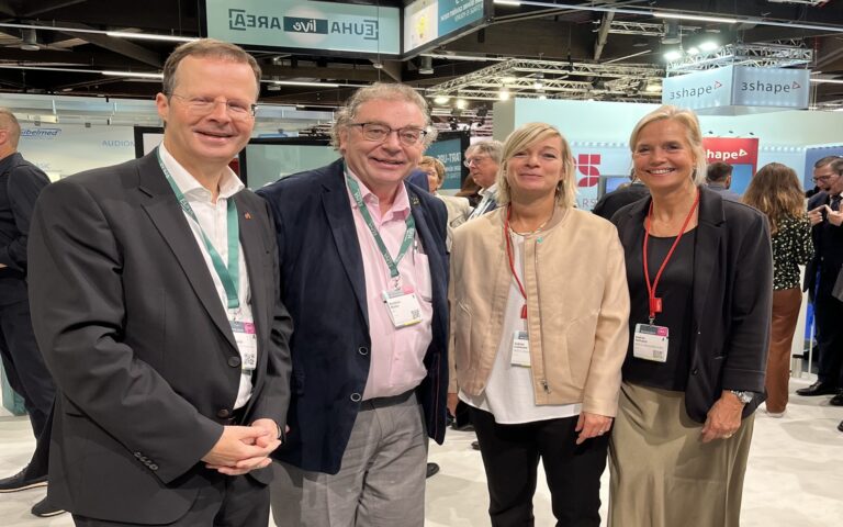 Vier Personen stehen in einer Messehalle, zwei Männer in Anzügen und zwei Frauen in Blusen und Jacken, alle mit Namensschildern an roten oder grünen Bändern um den Hals.