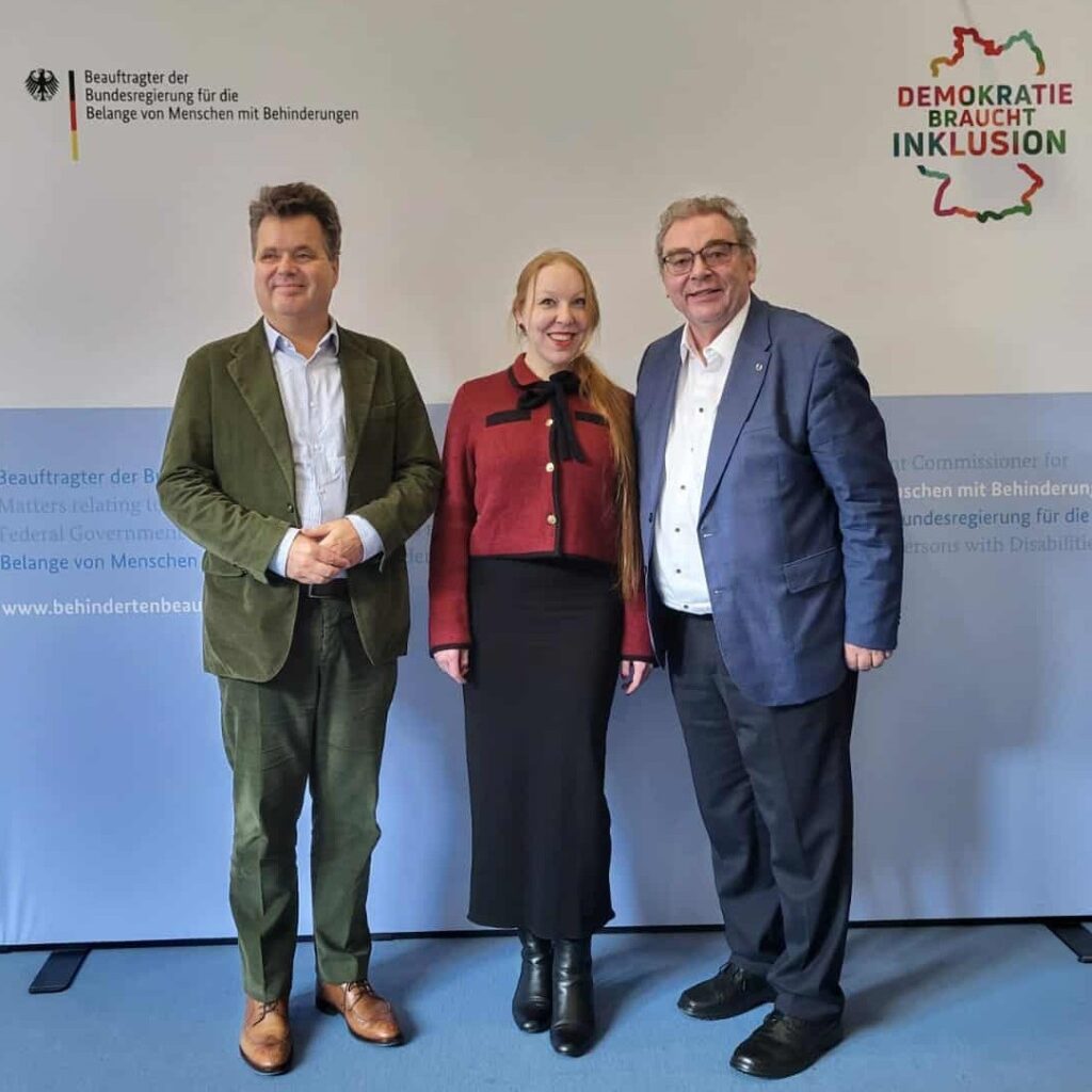 Drei Personen stehen nebeneinander vor einer Wand mit Text und einem Logo, zwei Männer tragen Anzüge, Frau trägt roten Blazer und schwarzen Rock.