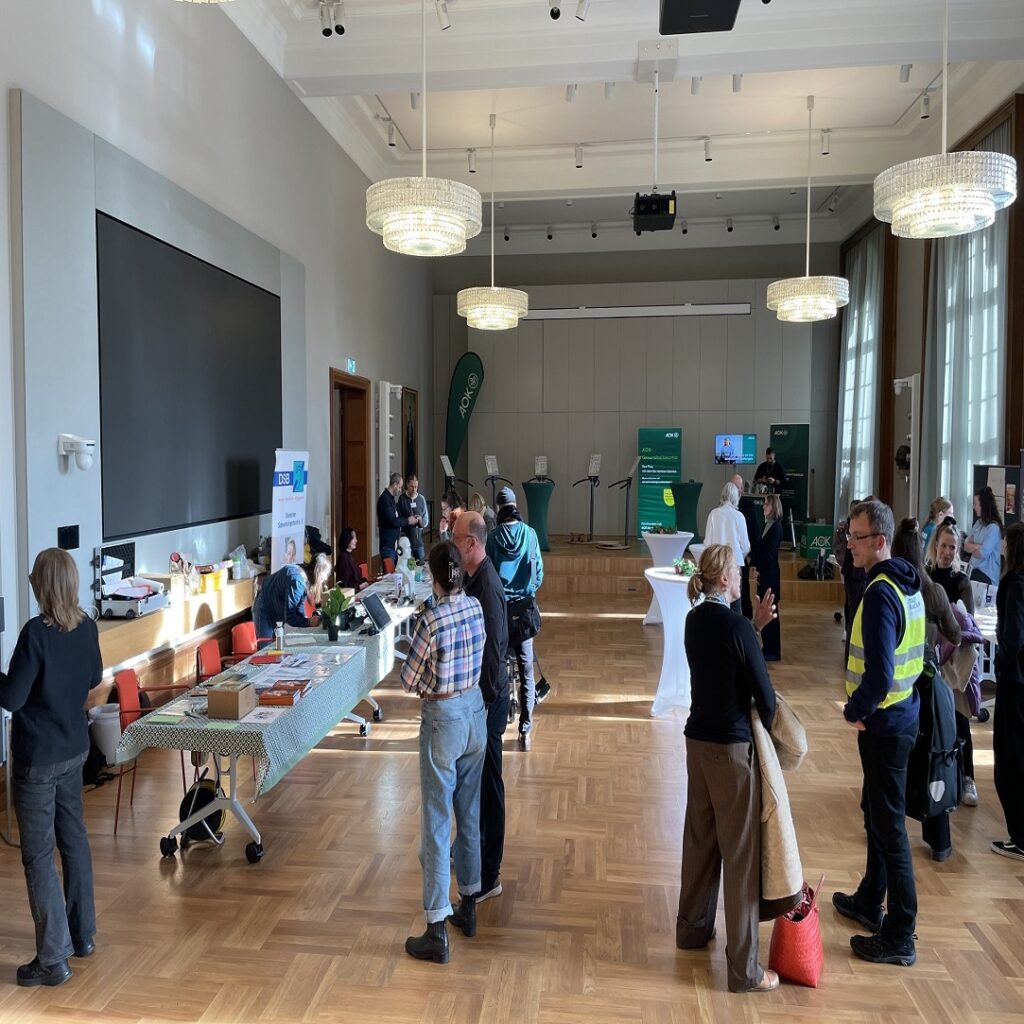 Großer heller Saal mit Parkettboden, mehreren Stehtischen und Menschen, die sich unterhalten und an einem langen Tisch mit Informationsmaterial stehen.