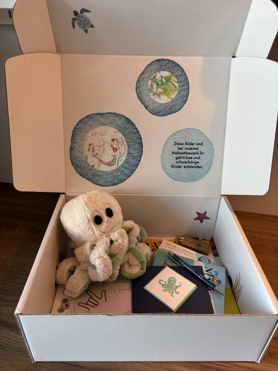 Offene weiße Box mit einem Plüsch-Oktopus, mehreren Karten und einer Innenseite mit drei runden Bildern und Text.