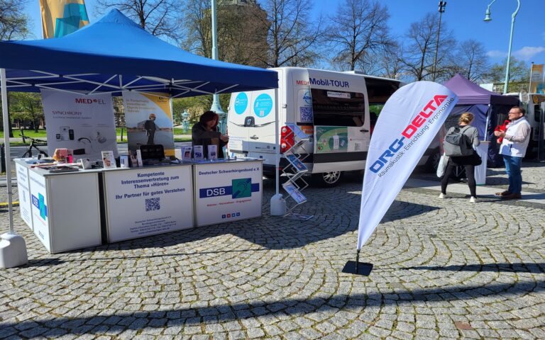Zwei Informationsstände mit blauen und violetten Zeltdächern, ein weißer Transporter mit Aufschrift 'HÖRMobil-TOUR'.