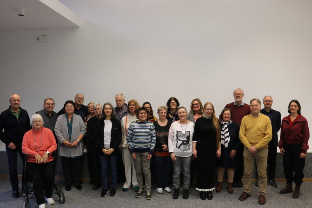 Gruppe von 22 Erwachsenen steht in zwei Reihen vor einer weißen Wand.