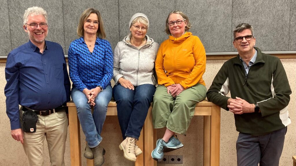 Fünf Personen, drei Frauen sitzen auf hohen Holzstühlen, zwei Männer stehen daneben, alle in legerer Kleidung vor grauer Wand.