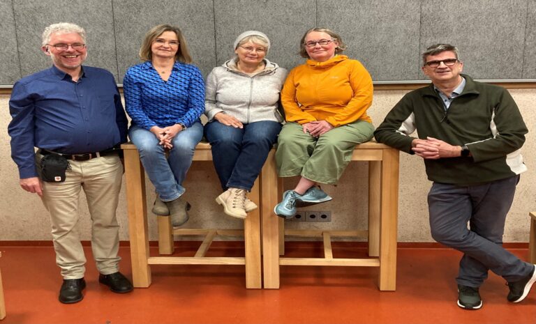 Fünf Personen, drei Frauen und zwei Männer, sitzen und lehnen an zwei hohen Holztischen in einem Raum mit grauer Wand.