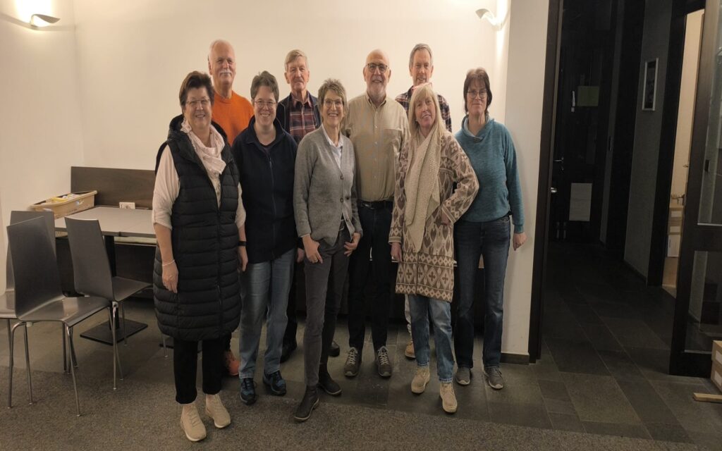 Gruppe von Menschen in einem Raum stehen vor einer weißen Wand mit Stühlen und Tischen.