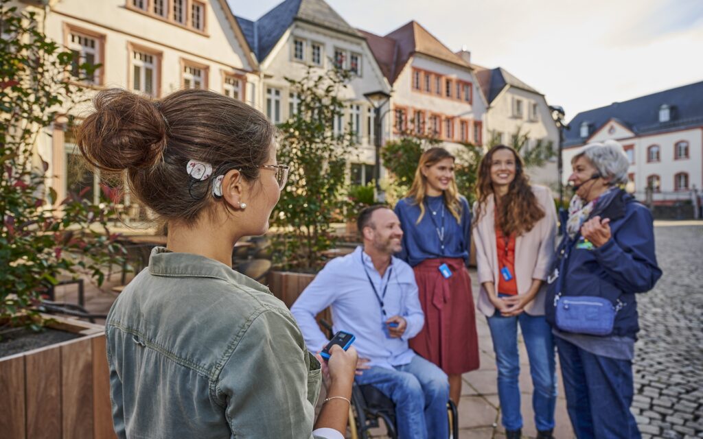Junge Frau mit Hochsteckfrisur und grünem Hemd hält ein Gerät in der Hand und steht vor einer Gruppe von vier Personen auf einem gepflasterten Platz mit historischen Gebäuden im Hintergrund.