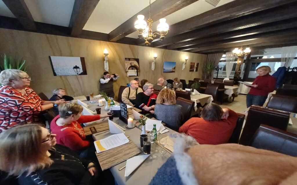 Gruppe von Menschen sitzt an zwei langen Tischen in einem rustikalen Raum mit Holzbalkendecke und Kronleuchtern, eine Person steht und spricht.