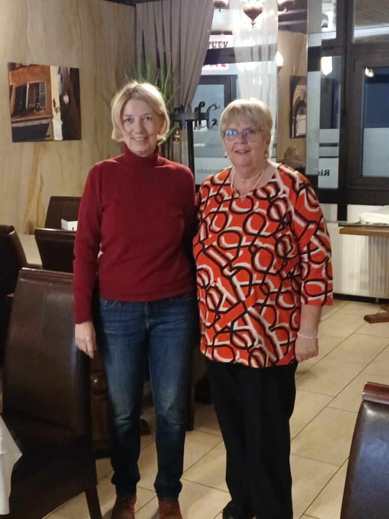 Zwei Frauen stehen nebeneinander in einem Raum mit Stühlen und einem Fenster im Hintergrund, eine trägt einen roten Pullover und Jeans, die andere ein gemustertes Oberteil mit schwarzen Hosen.