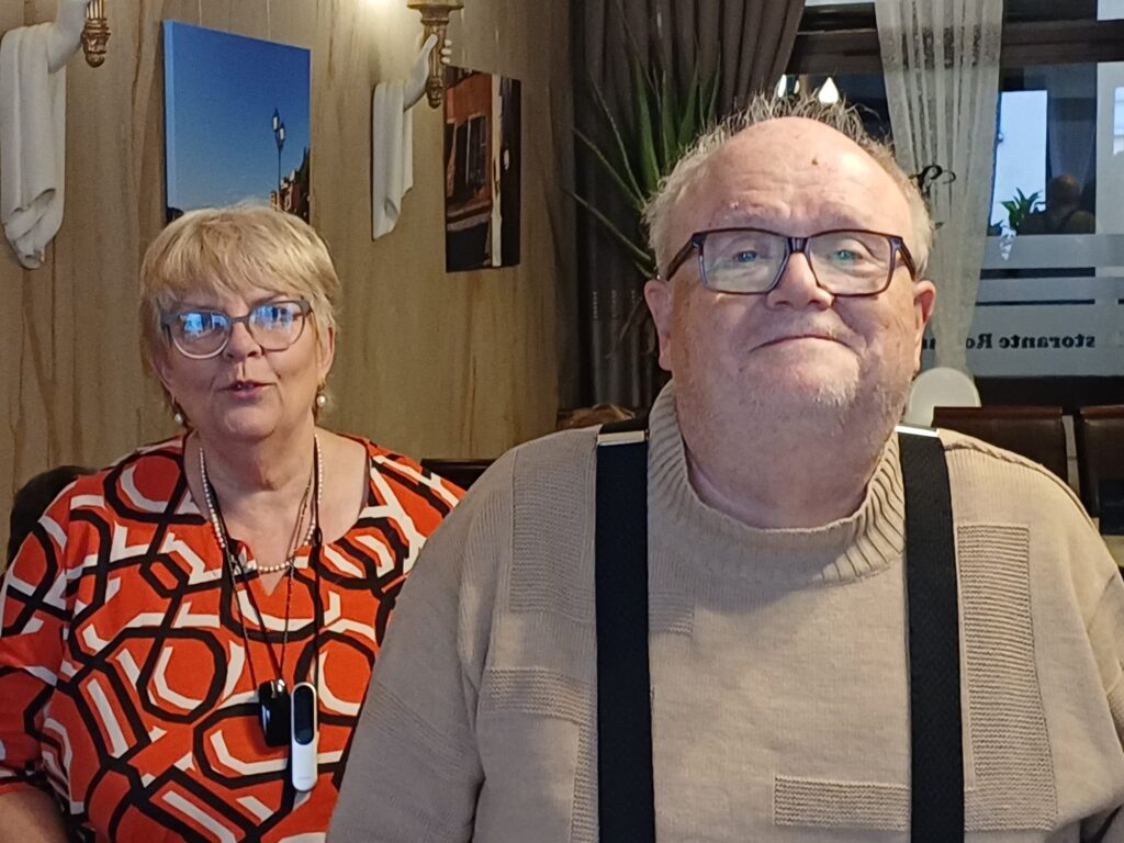 Zwei ältere Personen sitzen nebeneinander in einem Raum mit Holzwänden und dekorativen Bildern an der Wand