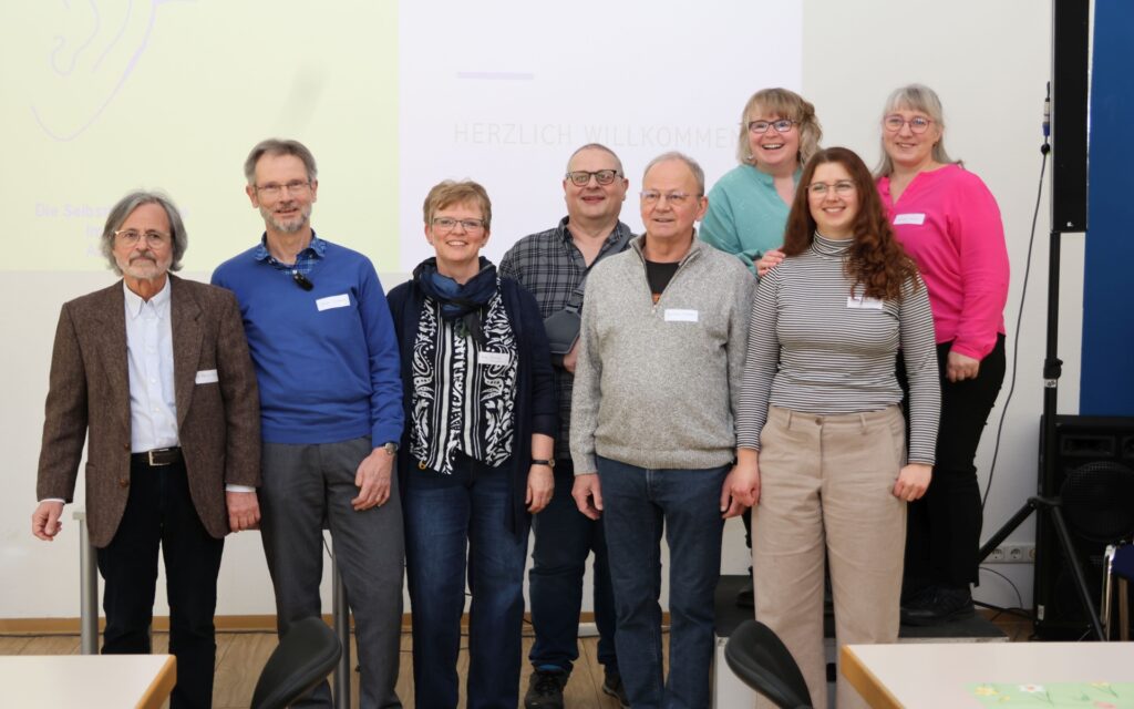 Gruppe von acht Personen steht in einem Raum vor einer weißen Wand zum Gruppenbild aufgestellt.