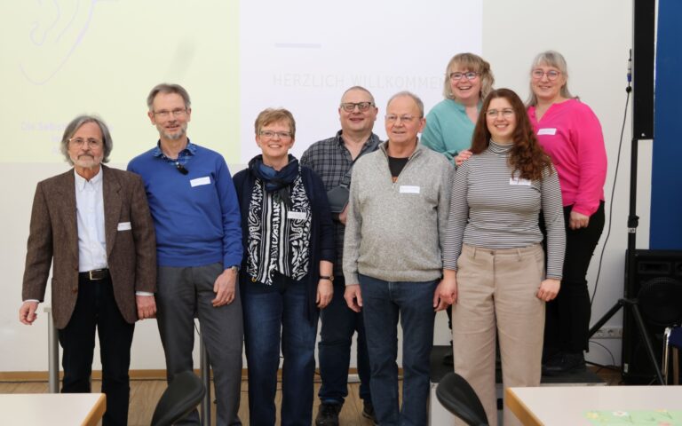 Gruppe von acht Personen steht in einem Raum vor einer weißen Wand zum Gruppenbild aufgestellt.