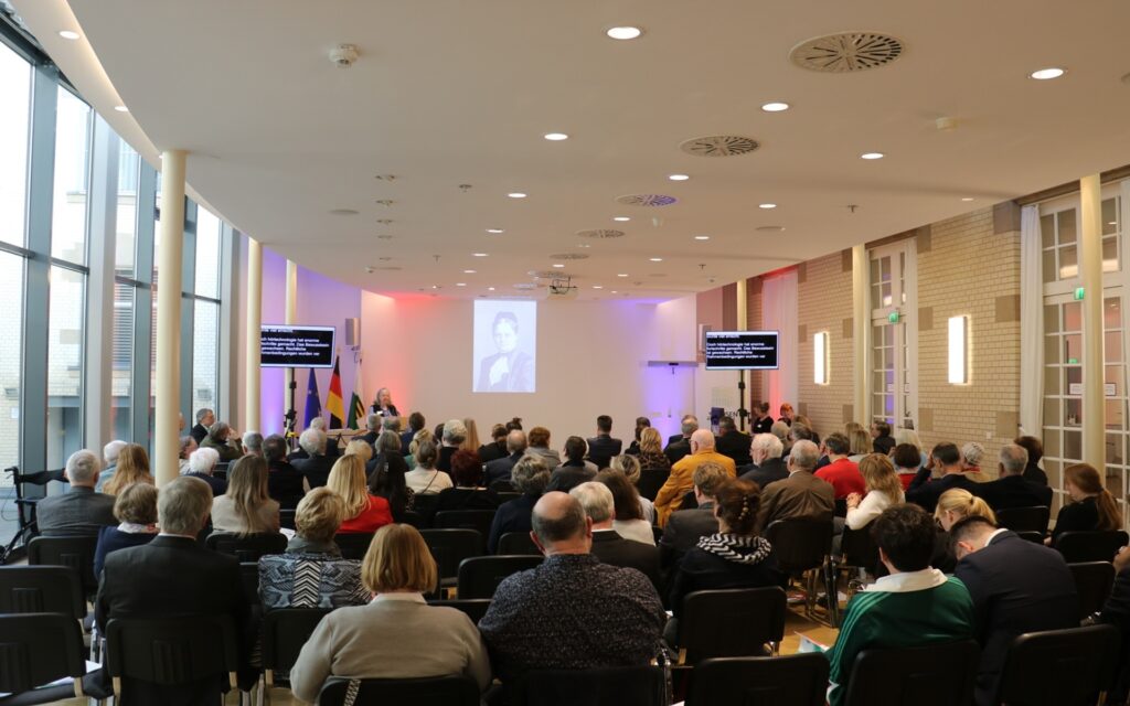 Publikum sitzt in einem hellen Konferenzraum mit großen Fenstern und blickt auf eine Leinwand und zwei Monitore mit Texten.