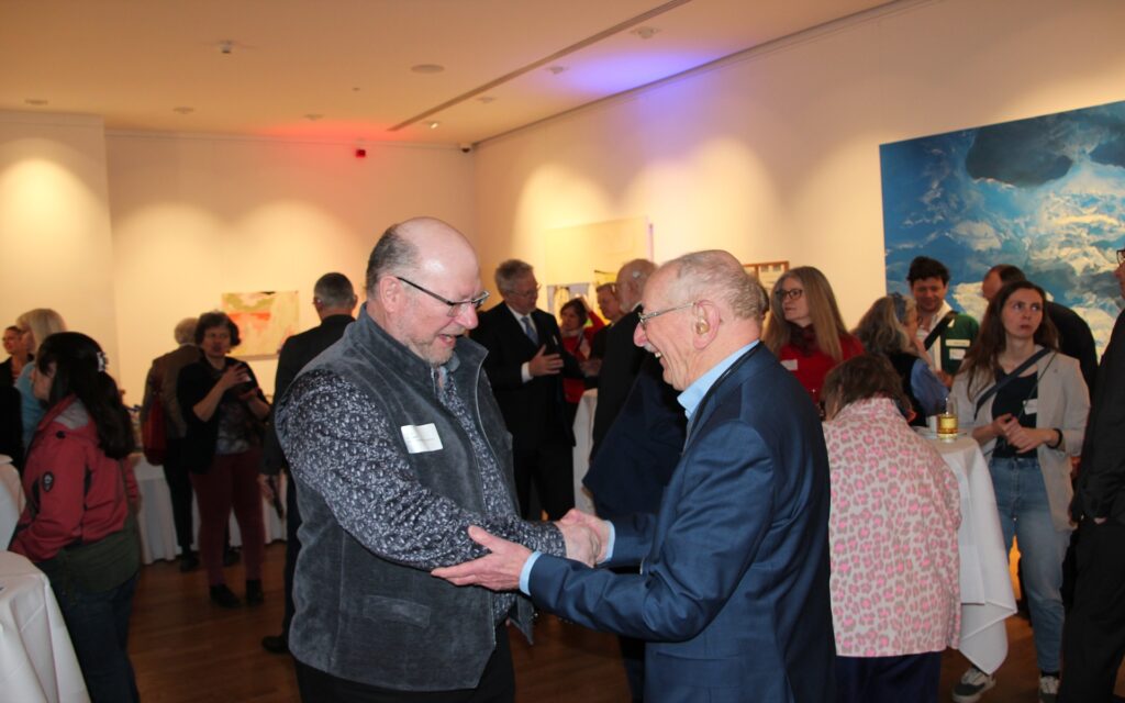 Zwei Männer im Vordergrund schütteln sich freudestrahlend die Hand in einem Raum mit mehreren Menschen und Kunstwerken an den Wänden.