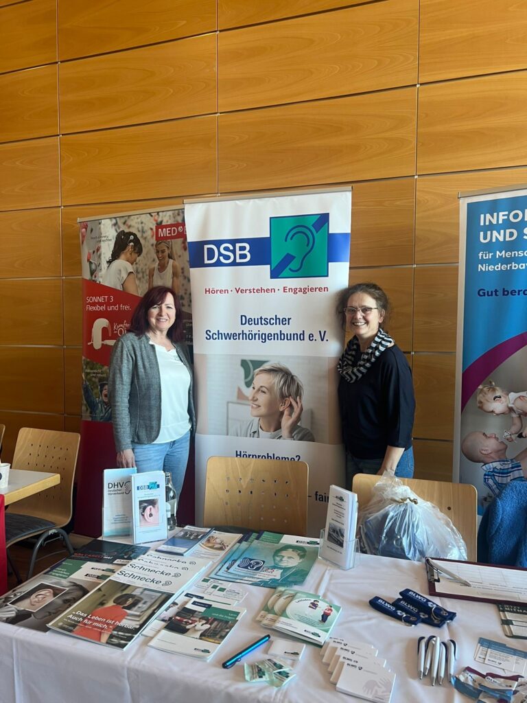 Zwei Frauen stehen an einem Infostand mit Flyern und Bannern des Deutschen Schwerhörigenbunds vor einer Holzpaneelwand.