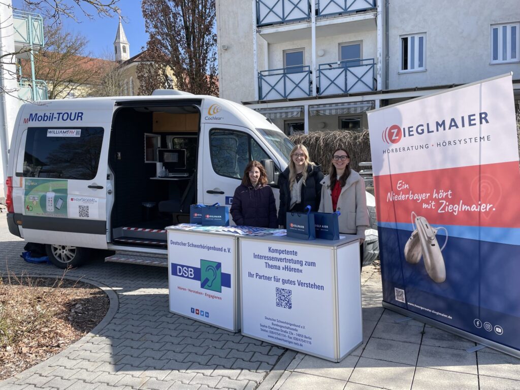 Drei Personen stehen vor einem weißen Van mit geöffnetem Seitentür und zwei Infoständen mit Bannern zu Hörsystemen und Schwerhörigenvertretung in einem sonnigen Innenhof mit Wohngebäuden im Hintergrund.