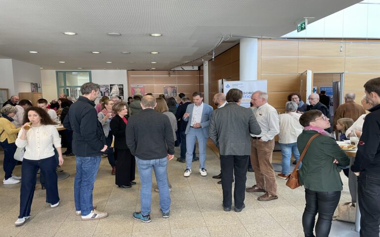 Gruppe von Menschen in einem hellen Raum mit Holzverkleidung, die sich unterhalten und Getränke halten.