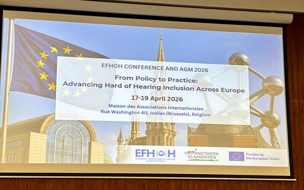 Projektionsleinwand mit Text zu einer Konferenz über Hörbehinderung in Europa vor einem Hintergrund mit dem Atomium in Brüssel und der Europaflagge.