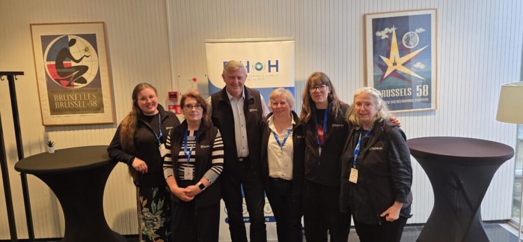 Sechs Personen stehen nebeneinander vor einem Roll-up mit dem Schriftzug 'EFHOH'. Sie tragen überwiegend dunkle Kleidung und Namensschilder. Die Gruppe steht in einem hellen Raum mit zwei Stehtischen, einer Stehlampe und zwei gerahmten Plakaten an der Wand.